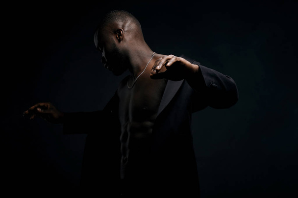 Dark key portrait of sexy dancer man topless, with jacket on shoulders and chainlet on neck. Studio shot, black background - Photo, Image