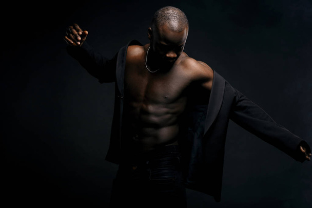 Dark key portrait of sexy dancer man topless, with jacket on shoulders and chainlet on neck. Studio shot, black background - Photo, image