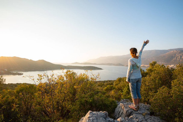 Happy traveler with open arms stands on the rock and admires turquoise waters of the Mediterranean Sea during sunset - adventure travel along Lycian way, Turkey. Back view, backlight. - Foto, Imagem