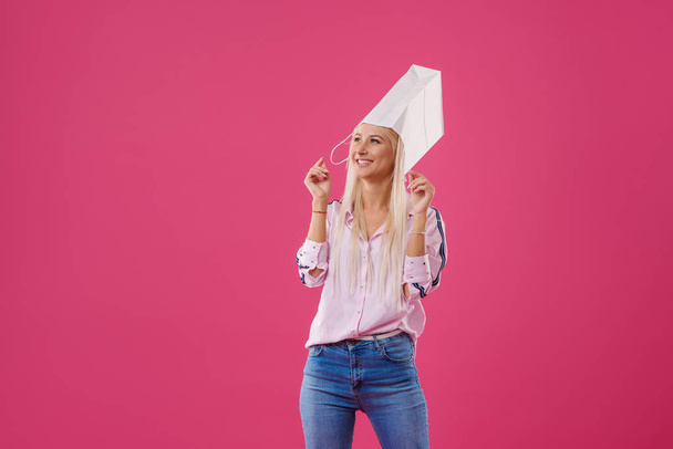 Portrait of an excited and fun blond girl with shopping bags on head over pink background - Фото, зображення