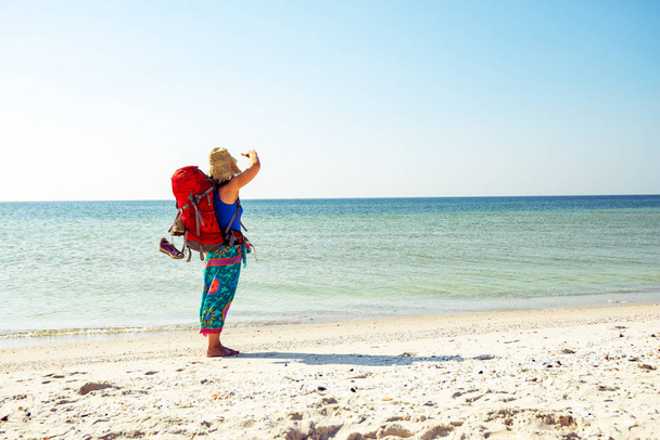 Woman in a pareo with backpack stands on the deserted beach next to the turquoise sea on a sunny day and looking into the distance. Awesome travel to the seashore. - Fotografie, Obrázek