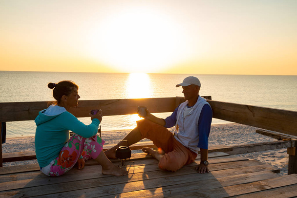 romantisches Paar entspannt sich auf einer alten hölzernen Bungalow-Veranda, trinkt Kaffee und genießt das Leben, während es den Sonnenuntergang über dem Meer bewundert. erstaunliche Reise an die Küste. Gegenlicht. - Foto, Bild