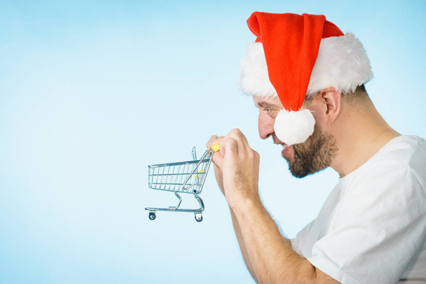 Man wearing santa claus hat holding empty shopping basket cart, on blue. Xmas, seasonal sales, boxing day concept. - Fotoğraf, Görsel