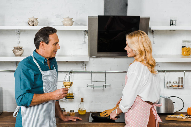 mature wife frying vegetables and husband holding glass of wine in kitchen - Photo, image