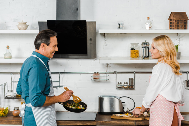 reife Frau schneidet Gemüse und Ehemann kocht auf Pfanne in Küche - Foto, Bild