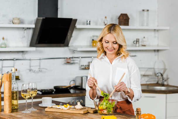 attractive smiling mature woman mixing salad in kitchen - Photo, Image