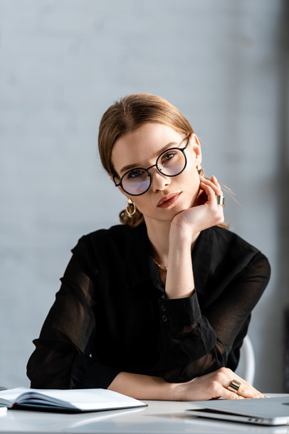 beautiful businesswoman in black clothes and glasses sitting on chair and looking at camera - Photo, Image
