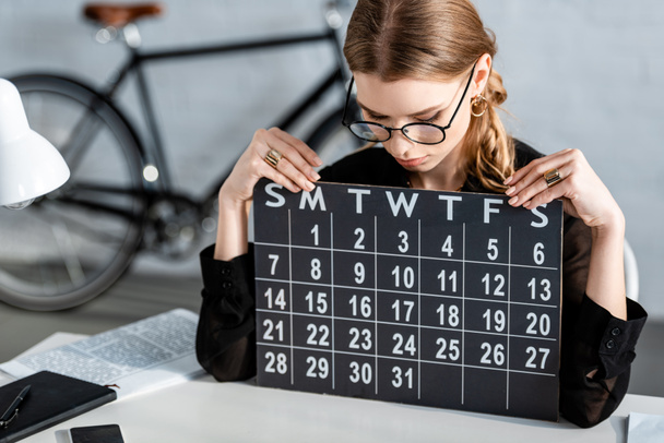 schöne Geschäftsfrau in schwarzer Kleidung und Brille sitzt auf einem Stuhl und schaut auf den Kalender - Foto, Bild
