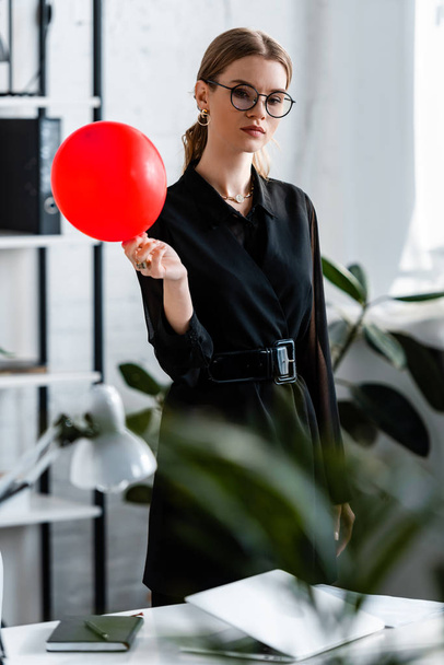 attraktive Frau in schwarzer Kleidung mit rotem Luftballon und Blick in die Kamera - Foto, Bild