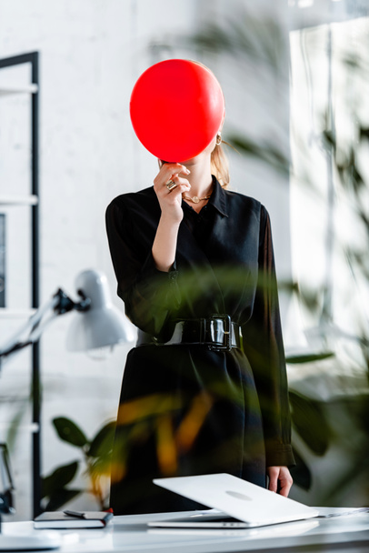 nő fekete ruhában közelében notebook, és elrejtik arc mögött piros léggömb  - Fotó, kép
