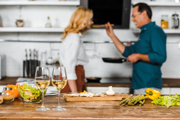 selective focus of mature husband feeding wife in kitchen, wineglasses on tabletop - Foto, Imagen
