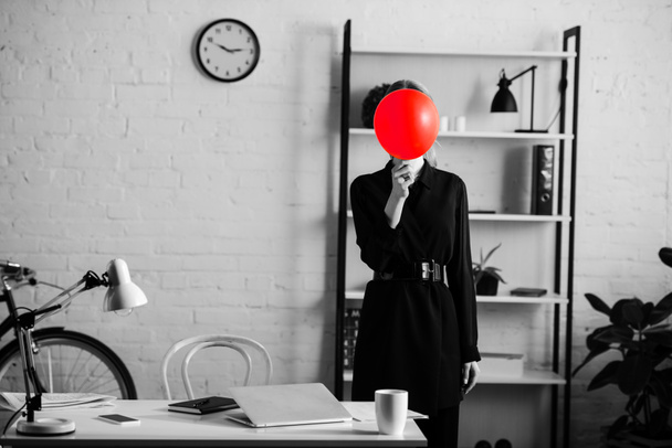 negro y blanco de mujer de negocios en ropa negra de pie cerca de la mesa y los estantes y ocultar la cara detrás de globo rojo
  - Foto, Imagen
