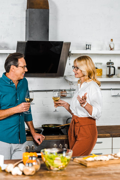 rijpe vrouw en man glazen wijn houden en kijken elkaar in de keuken - Foto, afbeelding