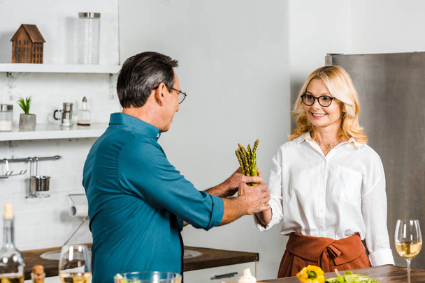 mature wife giving asparagus to husband for cooking in kitchen - 写真・画像