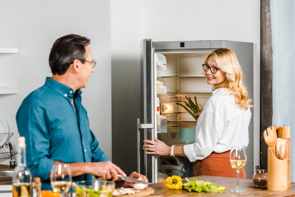 kypsä vaimo ottaa parsaa jääkaapista ja aviomies leikkaamalla vihanneksia keittiössä
 - Valokuva, kuva