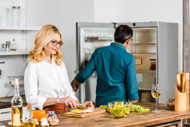 mature wife cutting vegetables and husband looking at fridge in kitchen - Foto, imagen