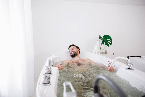 Pleased man turning his head while sitting in hydro massage bathtub - 写真・画像