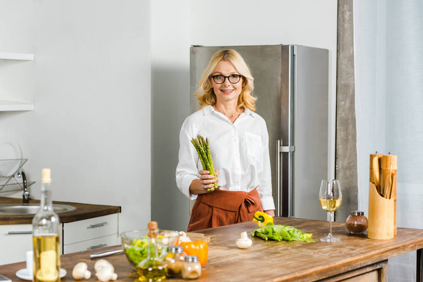 attractive mature woman holding asparagus in kitchen and looking at camera - Fotoğraf, Görsel