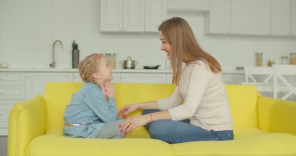 Positive excited little girl with pigtails and charming mother enjoying leisure together on sofa. Stunning mom and cheerful smiling elementary age girl communicating and bonding at home during weekend - Imágenes, Vídeo