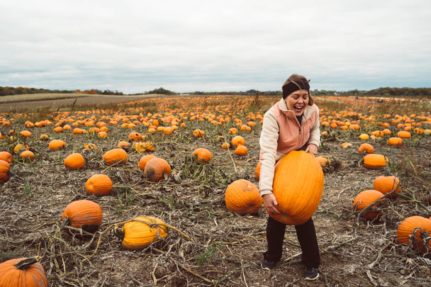 Adult woman (30s) attempts and struggles to lift and to pick up a giant pumpkin from a pumpkin patch. Smiling and laughing and having fun - Photo, Image