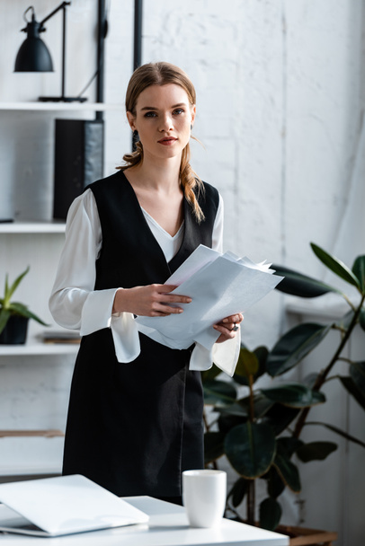 beautiful businesswoman in formal wear holding documents and looking at camera at workplace - Photo, Image