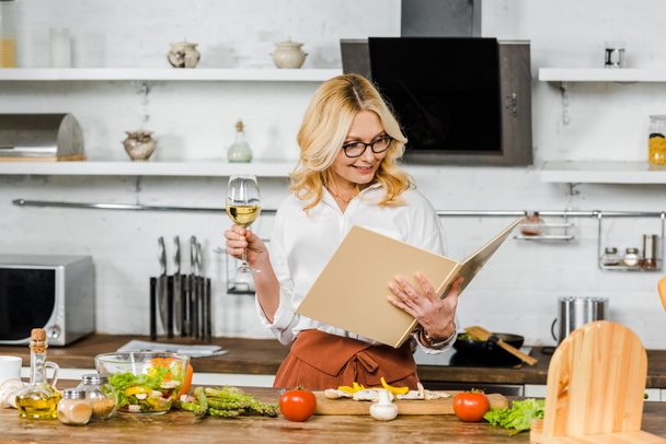 ελκυστική ώριμη γυναίκα κρατώντας το ποτήρι κρασί και να ψάχνει στο βιβλίο μαγειρικής στην κουζίνα - Φωτογραφία, εικόνα