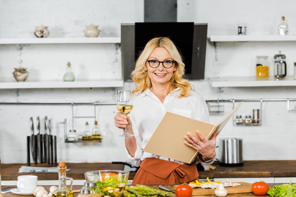 smiling attractive mature woman holding glass of wine and cookbook in kitchen - Foto, Imagem