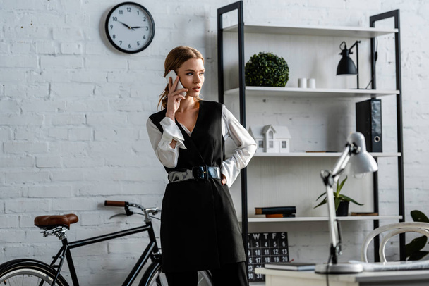 businesswoman in formal wear with hand on hip talking on smartphone in office - Photo, Image