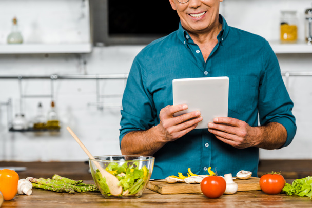 обрізане зображення усміхненого зрілого чоловіка, який використовує планшет під час приготування їжі на кухні
 - Фото, зображення