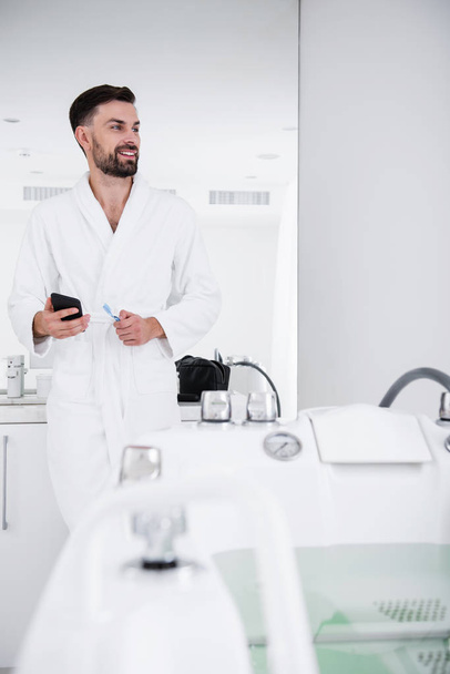Waist up of smiling man in white bathrobe standing in the bathroom - Фото, изображение