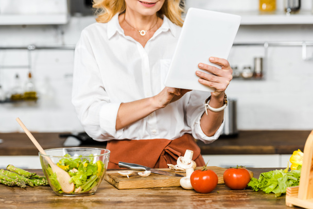 cropped image of mature woman reading recipe from tablet during cooking in kitchen - Фото, зображення