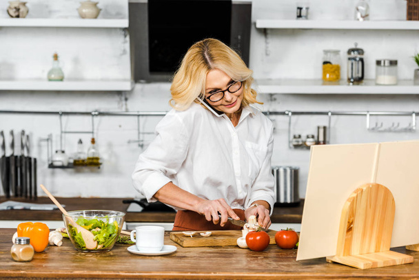 attrayant femme mature parler par smartphone, couper des légumes et regarder le livre de recettes dans la cuisine
 - Photo, image