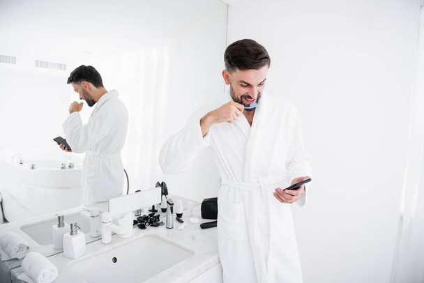Emotional man brushing his teeth and looking at smartphone - Foto, Imagen