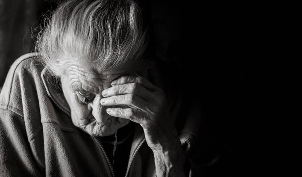 Black and white image of an old depressed woman. An elderly lonely woman sits at a table in the kitchen near the window and drinking tea. - Photo, Image