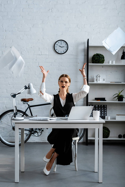 dissatisfied businesswoman in formal wear sitting at desk, gesturing with hands and throwing documents in air at workplace - Fotografie, Obrázek