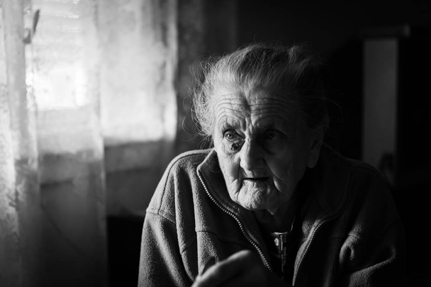 Imagem em preto e branco de uma velha mulher deprimida. Uma mulher idosa e solitária senta-se a uma mesa na cozinha perto da janela e bebe chá
. - Foto, Imagem