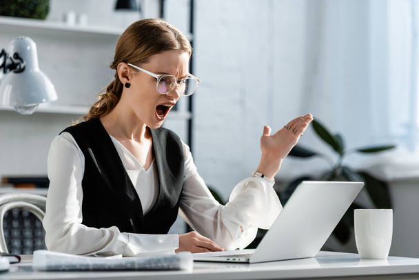 schockierte Geschäftsfrau in offizieller Kleidung, die am Computer-Schreibtisch sitzt und am Arbeitsplatz mit den Händen gestikuliert - Foto, Bild