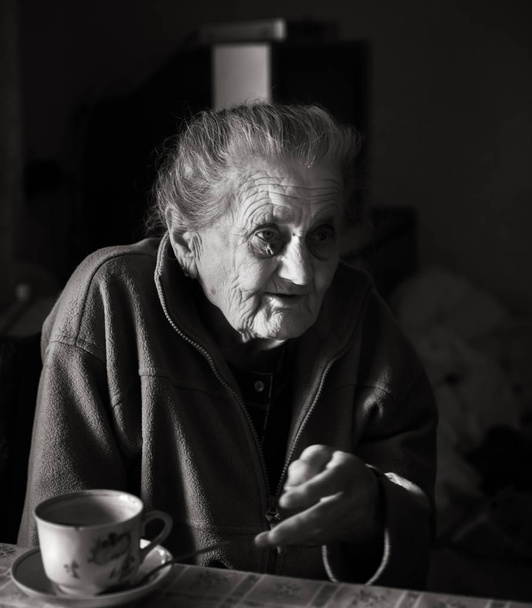 Imagem em preto e branco de uma velha mulher deprimida. Uma mulher idosa e solitária senta-se a uma mesa na cozinha perto da janela e bebe chá
. - Foto, Imagem