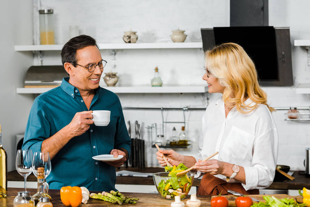 mature wife mixing salad and smiling husband drinking tea in kitchen - Foto, imagen