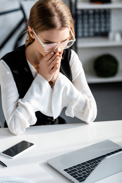 selektiver Fokus einer schockierten Geschäftsfrau in formeller Kleidung mit gefalteten Händen, die am Computertisch am Arbeitsplatz sitzt - Foto, Bild