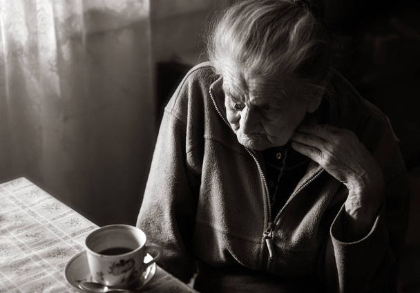 Zwart-wit beeld van een oude depressieve vrouw. Een eenzame bejaarde vrouw zit aan een tafel in de keuken in de buurt van het venster en drinken thee. - Foto, afbeelding