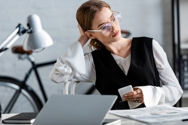 mujer de negocios en ropa formal y gafas sentado en el escritorio de la computadora y el uso de teléfonos inteligentes en el lugar de trabajo
 - Foto, Imagen