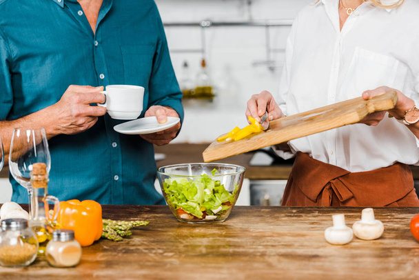imagem cortada de mulher madura colocando legumes na tigela e marido segurando xícara de chá na cozinha
 - Foto, Imagem