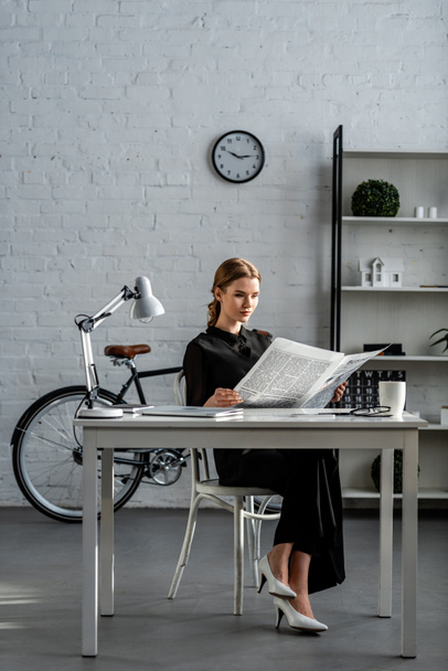 businesswoman in black formal wear sitting at desk and reading newspaper in office - Foto, afbeelding