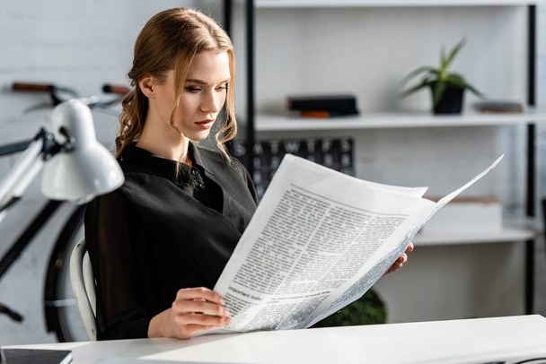 focused businesswoman in black formal wear sitting at desk and reading newspaper at workplace - Fotoğraf, Görsel