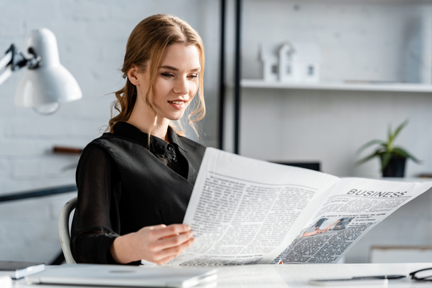 smiling businesswoman in black formal wear sitting at desk and reading newspaper at workplace - Фото, изображение