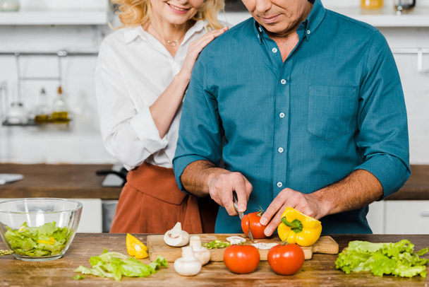 cropped image of mature wife hugging husband while he cooking salad in kitchen - Foto, imagen