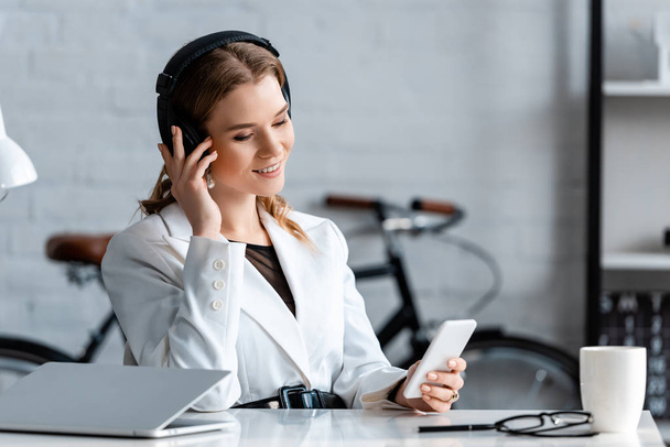 beautiful smiling businesswoman in headphones sitting at desk and using smartphone at workplace - Foto, Imagem