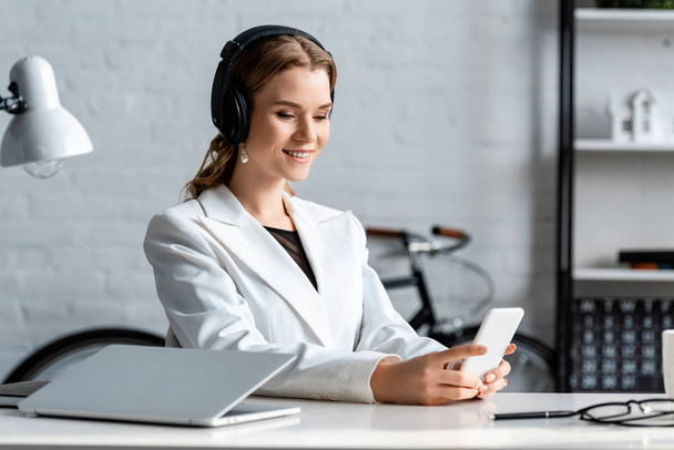 smiling businesswoman in headphones and formal wear sitting at desk and using smartphone at workplace - Photo, image