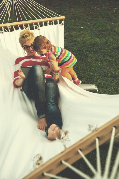 Happy mother and her little daughter enjoying free time using tablet computer while relaxing in a hammock during sunny day on holiday home garden - 写真・画像